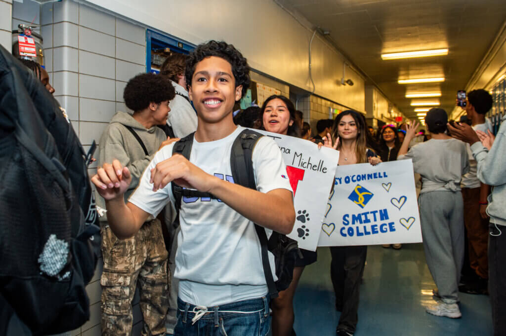 Students walk through a school hallway lined with cheering peers, some holding congratulatory signs for college acceptance, showcasing the student leadership networks mission to empower and celebrate student achievement.
