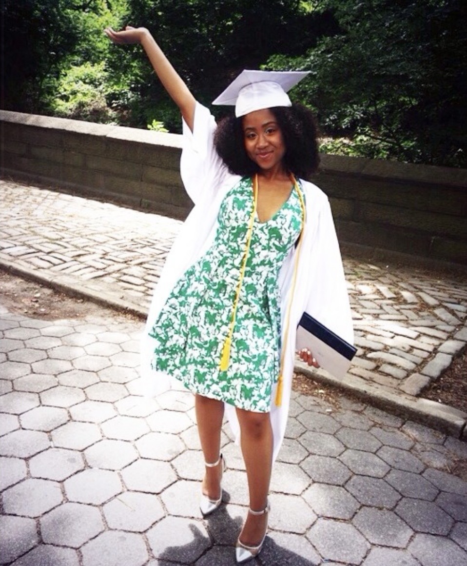 A smiling graduate in a white cap and gown over a green and white patterned dress, holding a diploma and waving.