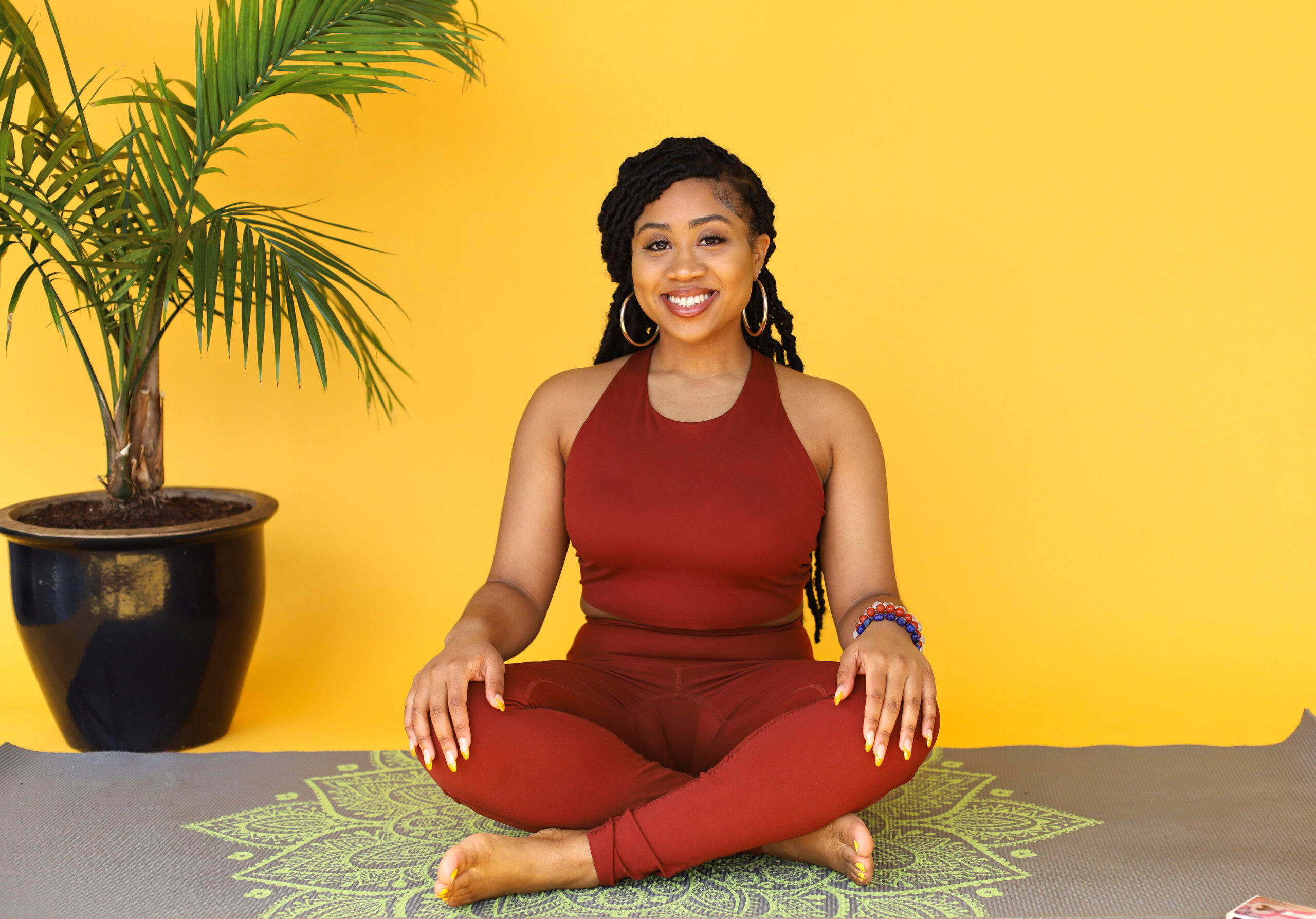 A woman smiling while sitting in a cross-legged yoga pose on a patterned mat next to a potted plant against a yellow background.